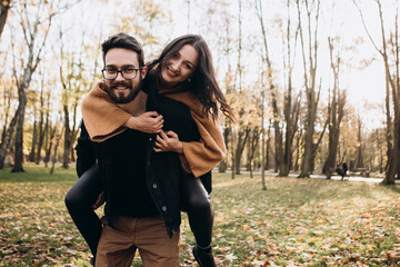Heterosexual caucasian young loving couple, husband carrying on back his wife in sunny weather, hugging smiling kissing laughing spending time together. Autumn, fall season, orange yellow red maple le