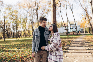 Fototapeta premium Heterosexual caucasian young loving couple walking outside in the city park in sunny weather, hugging smiling kissing laughing spending time together. Autumn, fall season, orange yellow red maple leav
