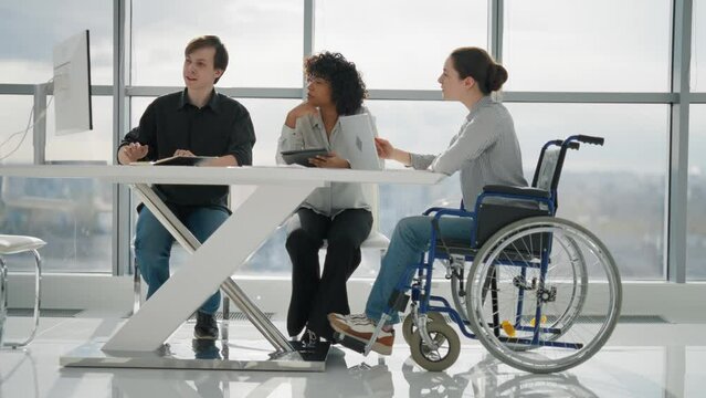 Paralyzed woman works with colleagues. Female in wheelchair has spine legs trauma discussing work project in office on meeting with multiethnic team. Mutual help, brainstorming, special needs person.