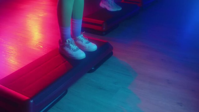 Cropped shot of legs of people from aerobics group doing steps on fitness equipment while working out in studio with neon lights