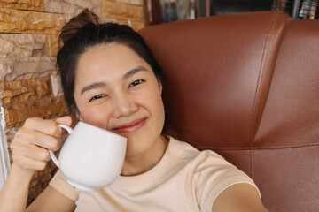 Close up of woman take selfie while holding white coffee mug, happy smiling to camera while drinking milk tea cup, happy sitting on couch at home alone.