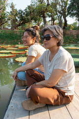 Side view of asian Thai Chinese elder mother and daughter sitting on the bank by lotus flower lake local lagoon, both woman happy smiling, travel on vacation.