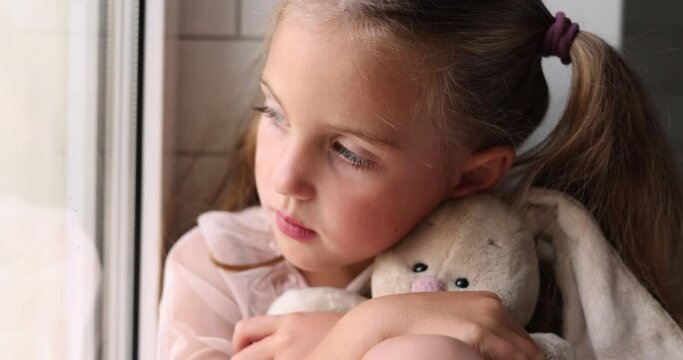 Close-up portrait of a little girl hugging a toy fluffy rabbit looking sadly out the window sitting on the windowsill waiting and feeling sad