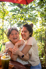 Asian Thai daughter hugging her elder mother, both woman playful and laughing together while sitting with cold drinks at cafe garden.