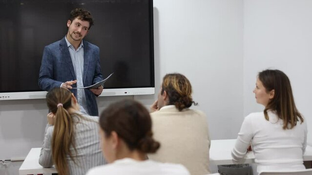 Young male teacher near interactive board, gesturing with hand while consecrating topic of lesson for adults in classroom