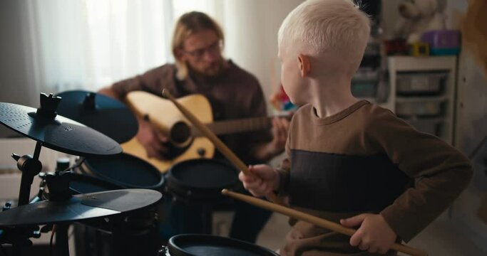 A happy albino boy with white hair plays on a special electronic drum kit using yellow drum sticks and his father, a blond man with long hair with a beard and glasses, plays the guitar with his son