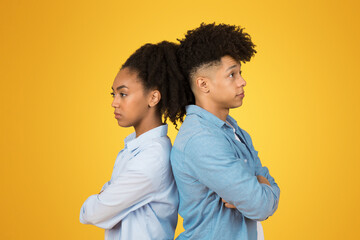 A teenage girl and boy stand back-to-back with arms crossed and displeased expressions, against a...