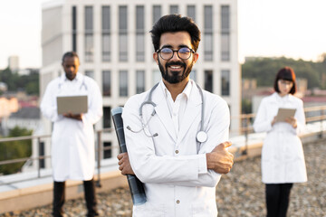 Portrait of confident young male doctor in white lab coat posing on camera with crossed hands and smiling. International colleagues of mixed nationality standing behind and operating modern gadgets.
