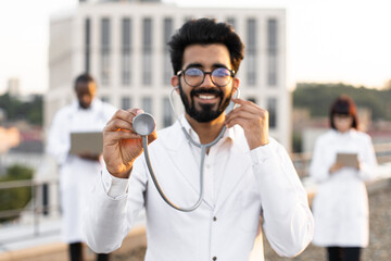Portrait of confident bearded male doctor in white lab coat posing on camera holding stethoscope. International colleagues of mixed nationality standing behind and operating modern gadgets.