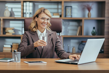 Fototapeta premium Smiling mature businesswoman with glasses working on laptop in home office, holding a glass of water reflecting a healthy lifestyle.