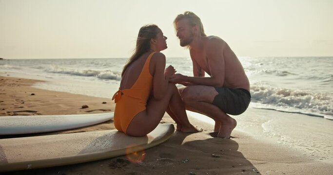 A blonde girl in an orange swimsuit sits on a surfboard, communicates with her blonde boyfriend and kisses him on the sandy beach near the sea in the morning at Sunrise