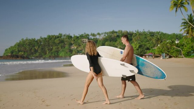 Athletic fit man, slender woman walk to ocean beach, carry surfboards. Young surfers couple dating. Boyfriend, girlfriend go surfing on sandy sea shore. Slim girl, guy with boards. Paradise coastline.