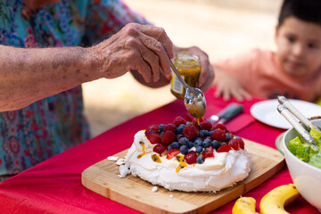 little kid out of focus looks on as older person spoons passionfruit over pavlova laden with berries