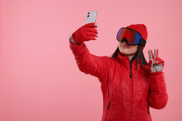 Woman in winter sportswear and goggles taking selfie and showing peace sign on pink background....
