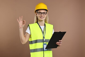 Engineer in hard hat holding clipboard and showing ok gesture on brown background