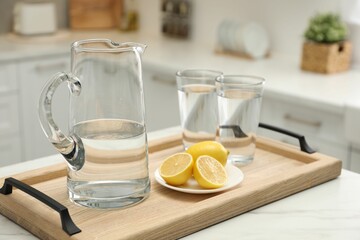 Jug, glasses with clear water and lemons on white table in kitchen