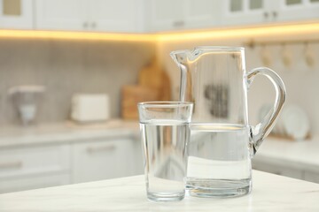 Jug and glass with clear water on white table in kitchen, space for text