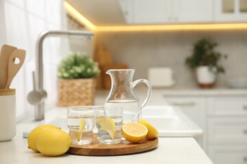 Jug, glasses with clear water and lemons on white table in kitchen, space for text