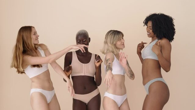 Happy gen z multiethnic girls wearing underwear dancing together on beige background. Four pretty diverse young women, smiling multicultural ladies models group having fun.