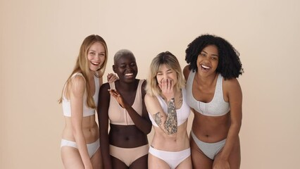 Happy diverse different gen z young women group wearing underwear standing together having fun on beige background. Four positive multiethnic girls beauty models hugging laughing looking at camera.
