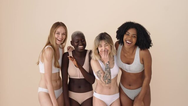 Happy diverse different gen z young women group wearing underwear standing together having fun on beige background. Four positive multiethnic girls beauty models hugging laughing looking at camera.