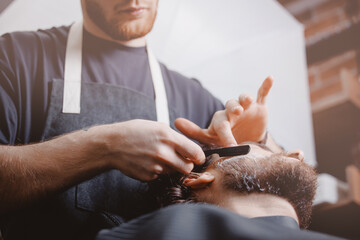 Hipster man sitting in armchair barber shop, hairdresser shaves men beard with dangerous razor