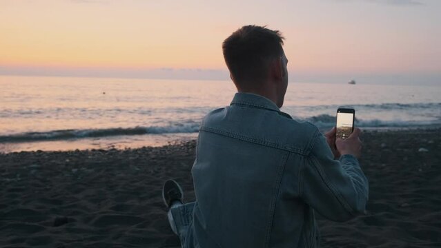 Back view of man tourist shooting video, taking panoramic photo of sea sunset on black sand beach using smartphone. Male with mobile phone takes pictures, enjoys nature, freedom, relaxing