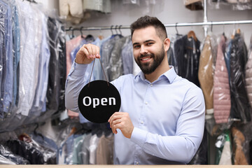 Dry-cleaning service. Happy worker holding Open sign at counter indoors