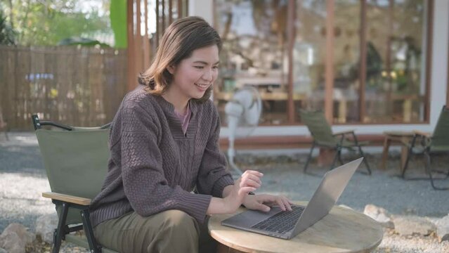 Young happy smiling Asain woman in cafe working video call facetime type project working on her laptop notebook personal computer, Female begins typing say hello on screen
