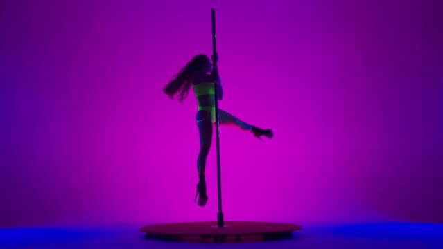 Young spectacular woman shows her flexibility and plasticity during pole dancing. Woman in yellow net suit dancing on pylon in studio on pink background with circular light.