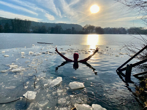 Eisbaden, Frau schwimmt im Eiswasser und h&auml;lt die Arme in der Siegespose hoch, See zugefroren. Sonnenuntergang