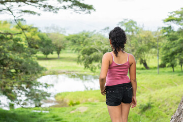 latina brunette girl standing with her back turned, in a paddock looking out over a lake in the distance