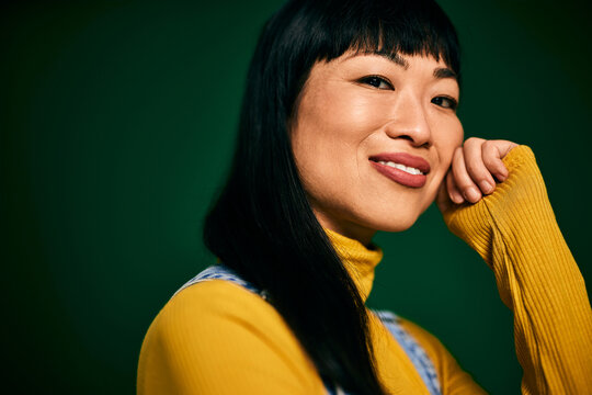 A happy Asian woman, smiling for the camera, dressed in a yellow sweater, has black hair.