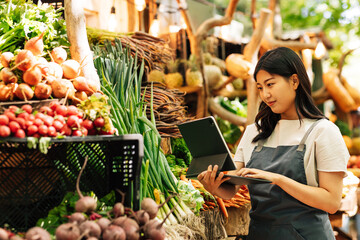 Young businesswoman in an apron working at a local food market. Female with digital tablet at a stall with vegetables.