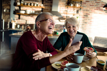 Two senior women having breakfast together and using smartphone at home