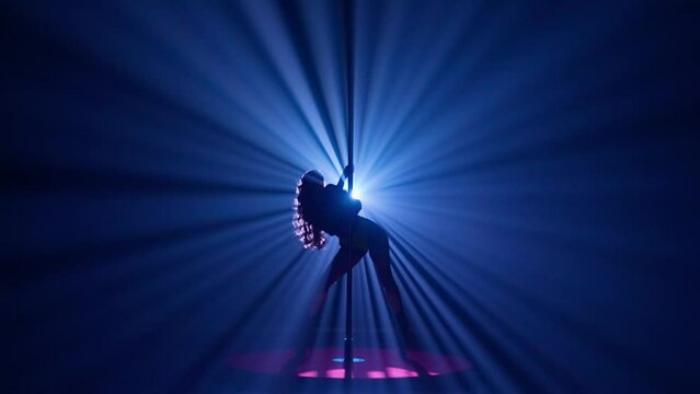 Dark silhouette of a woman spinning upside down on a pylon. Woman dancing on a pole in a dark studio against a bright spotlight.