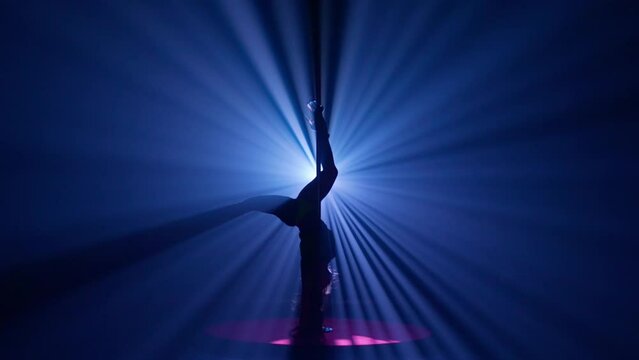 Dark silhouette of flexible and plastic woman dancing on a pole. Woman standing on her arms with her head down. Woman dancing on a pylon in a dark studio against a bright spotlight.