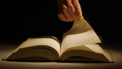 Studio shot of old retro hardcover book on dark background in warm light. Man holding page of old vintage book laying opened.