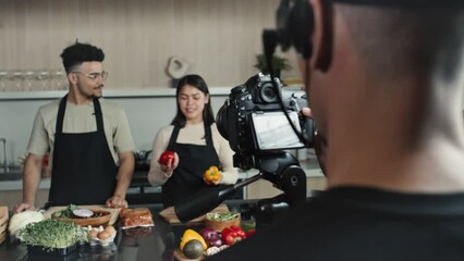 Over the shoulder shot of cameraman filming two young chefs standing behind kitchen table and telling about food ingredients while hosting TV culinary show