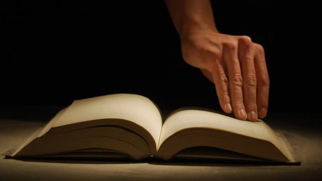 Close up studio shot of old retro hardcover book on dark background in warm light. Man comes and turns dusty page of old book.