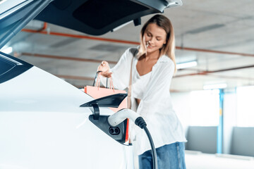 Young woman travel with EV electric car to shopping center parking lot charging in downtown city...