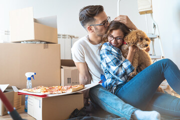 Young couple with a dog happy in their new family home sitting on the floor having pizza surrounded...