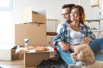 Portrait of happy smiling couple looking away and having pizza for lunch in new apartment together with poodle dog.