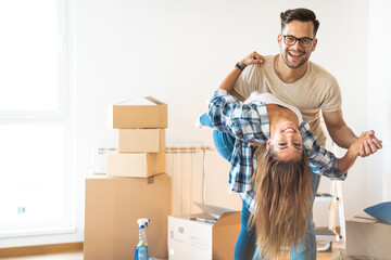 Cheerful smiling young couple looking at camera while dancing in new apartment with cardboard boxes...