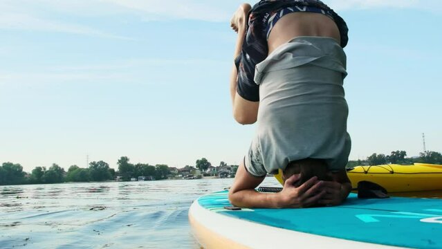 Sportsman practices yoga on sup board to alleviate stress and anxiety. Man does headstand to help build strength during summer vacation