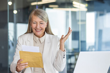 Happy gray-haired senior business woman is sitting in the office at the desk and is happy reading the received letter in the envelope.