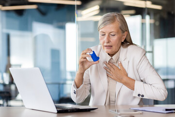 Senior gray-haired businesswoman sitting in the office at the desk and holding her hand to her...