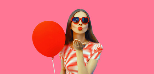 Portrait of happy young woman with red balloon blowing her lips wearing heart shaped sunglasses on pink studio background