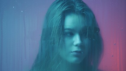 Portrait of female in neon light behind the glass window in steam and water drops. Girl with makeup looks around at camera through wet glass.