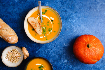 Pumpkin cream soup on blue background decorated with pumpkin, pumpkin seeds and fresh bread. Top overlay view. Autumn comfort food concept.